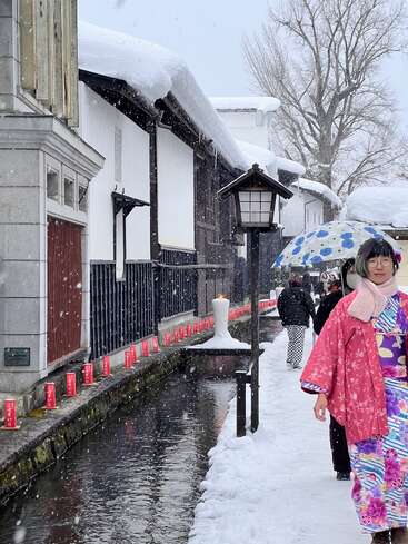 In einer traditionellen japanischen Straße fällt sanft Schnee. Menschen in farbenfrohen Kimonos und Regenschirmen spazieren an einem Kanal entlang, rote Laternen säumen den verschneiten Weg, und Laternen leuchten warm.