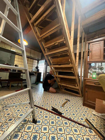 Eine Person baut oder repariert eine Holztreppe in einem Raum mit gemustertem Fliesenboden, umgeben von Werkzeug, einer Leiter und Möbeln im Vintage-Stil.