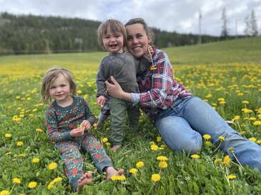Una mujer y dos niños se sientan descalzos en un campo de flores amarillas, sonriendo alegremente, rodeados de hierba verde y colinas bajo un cielo nublado.