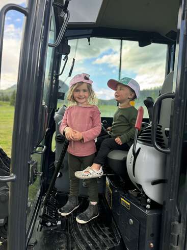 Duas crianças pequenas dentro de um veículo grande, possivelmente um trator. A menina está de pé e sorrindo; o menino está sentado ao seu lado. Ambos usam chapéus e roupas casuais.