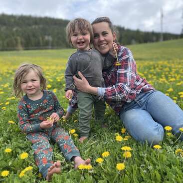 Eine Frau und zwei Kinder sitzen barfuß in einem gelben Blumenfeld und lächeln fröhlich, umgeben von grünem Gras und Hügeln unter einem wolkenverhangenen Himmel.