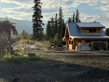 Uma pequena cabana de madeira fica sob um céu claro, cercada por árvores altas, aspersores regando o jardim e sacos de suprimentos empilhados ao lado da casa.