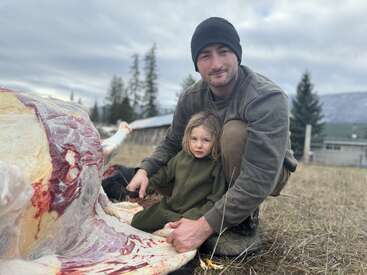 Um homem e uma jovem, ambos vestidos com roupas quentes, ajoelham-se ao lado de uma grande carcaça de animal ao ar livre, cercados por um cenário rural e céu nublado.