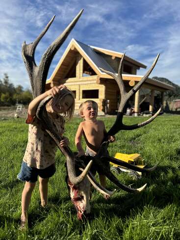 Duas crianças pequenas estão na grama verde em frente a uma cabana de madeira, segurando um crânio de veado grande e ensanguentado com chifres. Há um caminhão de brinquedo por perto.