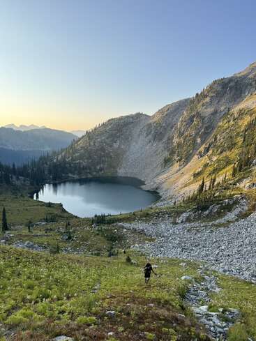 Um caminhante solitário desce uma encosta gramada em direção a um lago alpino sereno, cercado por montanhas rochosas, vegetação exuberante e um céu azul claro pela manhã.
