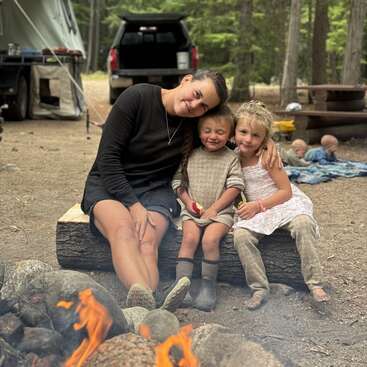A woman and two children sit closely on a log by a campfire, smiling. Tents, a truck, and more children are visible in the forested background.