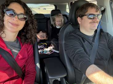 A family of four is enjoying a car ride. The parents sit in front smiling, while two children are in the back, one asleep, one awake.