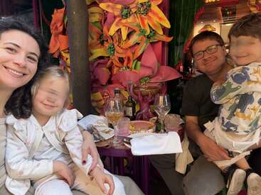 A smiling family of four enjoys a meal at a colorful restaurant decorated with large, vibrant flowers. Two children sit happily with their parents.