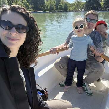A family of four enjoys a sunny day on a boat ride. Everyone is wearing sunglasses, smiling, and surrounded by peaceful water and green trees.
