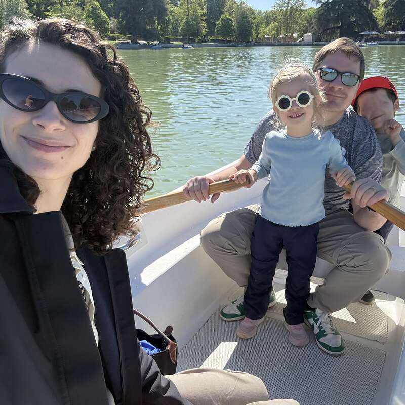 A family of four enjoys a sunny day on a boat ride. Everyone is wearing sunglasses, smiling, and surrounded by peaceful water and green trees.