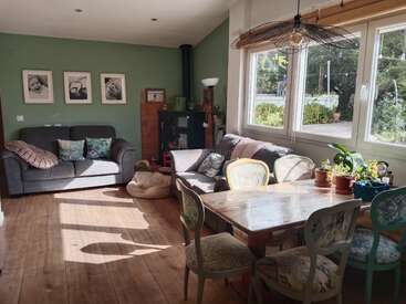Un salon confortable et ensoleillé comprend deux canapés gris, une table de salle à manger en bois, des chaises assorties, un poêle à bois, des murs verts, des plantes d'intérieur, de grandes fenêtres et des photos encadrées.