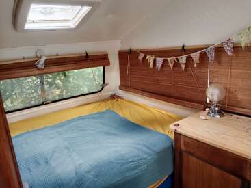 Cette chambre de camping confortable est dotée d'une lucarne, d'une tapisserie colorée, d'une literie bleue et jaune, de stores en bois, d'une lampe et d'une petite armoire en bois pour le rangement.