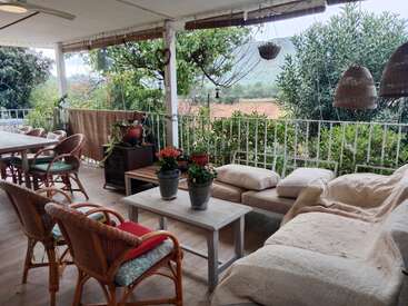 Un patio couvert confortable avec des chaises en osier, un canapé rembourré, des plantes en pot sur les tables, un décor rustique, des paniers suspendus, entouré d'une verdure luxuriante et d'une vue pittoresque sur les montagnes.