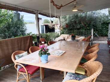Un patio extérieur confortable avec une grande table en bois, des chaises en osier, des fleurs en pot, un coin salon confortable, des lampes suspendues, un ventilateur de plafond, le tout entouré d'une végétation luxuriante.