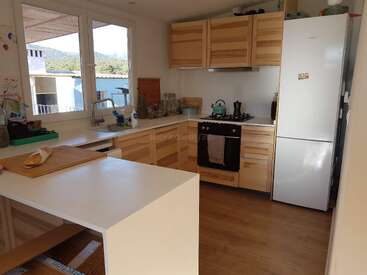 La cuisine moderne et lumineuse se compose d'armoires en bois clair, de comptoirs blancs, d'un grand réfrigérateur, d'un four, d'un îlot central, de grandes fenêtres et d'appareils électroménagers soignés, créant ainsi une atmosphère chaleureuse.