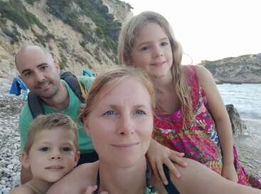 L'image montre une famille de quatre personnes posant pour un selfie sur une plage rocheuse, avec une falaise et des arbres en arrière-plan.