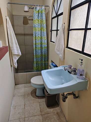 Este cuarto de baño tiene un lavabo azul, inodoro a juego, ducha con cortina estampada, dos toallas, ventanas esmeriladas, paredes alicatadas, dispensador de jabón, papelera y artículos de aseo.
