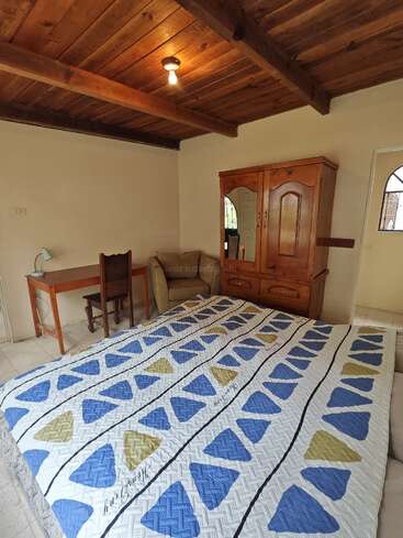 Este acogedor dormitorio cuenta con un techo de madera, una sola bombilla, una colcha estampada, un armario de madera con espejo y un escritorio con lámpara.