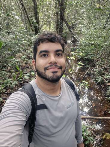 A man takes a selfie in a lush, green forest. He is wearing a gray shirt and backpack, standing near a small, reflective stream.