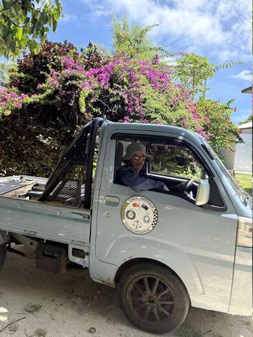 Ein Mann mit Hut sitzt in einem kleinen Pick-up, umgeben von üppigem Grün und leuchtend rosa Blumen, unter einem strahlend blauen Himmel.