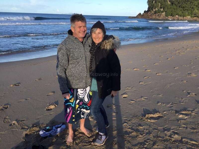 Das Bild zeigt einen Mann und eine Frau, die an einem Sandstrand stehen und sich umarmen, während im Hintergrund das Meer und die Wellen zu sehen sind.