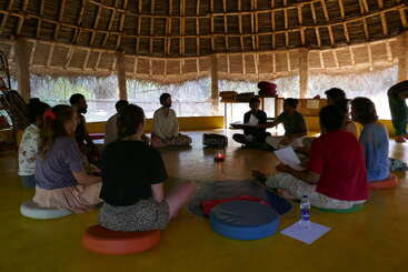 The image depicts a group of people seated in a circle, engaged in a discussion or meeting, with a table and chairs visible in the background, conveying a sense of community and collaboration.
