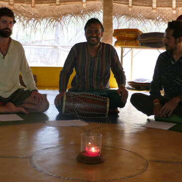The image depicts three men seated on the floor, surrounded by a candle and papers, with a drum positioned between them, in a room with a thatched roof and a window in the background.