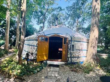 A cozy white yurt with bright yellow doors stands among tall trees, surrounded by greenery and pebbled paths, creating a peaceful and inviting woodland retreat.