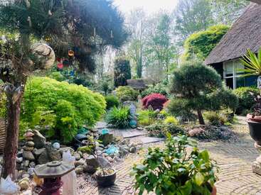 A serene garden with lush greenery, decorative rocks, a Buddha statue, vibrant plants, a thatched-roof house, and handcrafted ornaments, evoking a peaceful, Zen-like atmosphere.