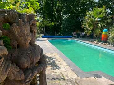 A stone statue stands beside a serene swimming pool, surrounded by lush greenery, palm trees, patio chairs, and a colorful sculpture under bright sunlight.