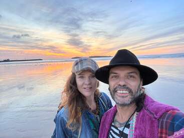 A smiling couple takes a selfie on a beach at sunset. The sky is colorful with orange, pink, and blue hues, reflecting on the wet sand.
