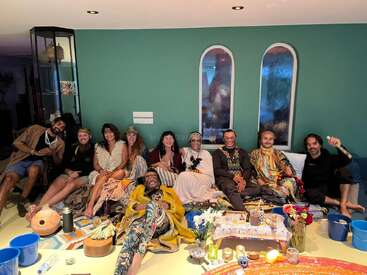 A joyful group of eleven people sits together indoors on blankets and cushions, surrounded by candles, flowers, musical instruments, and spiritual or ceremonial items, smiling warmly.