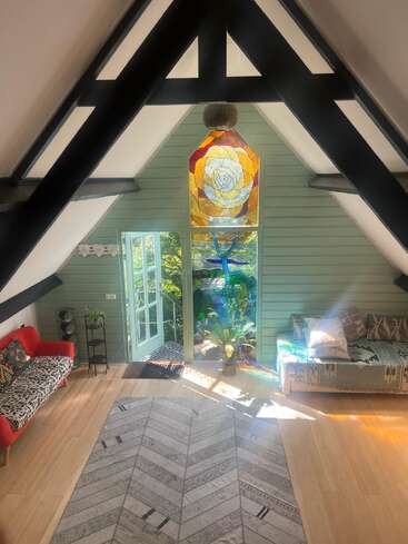 A cozy, sunlit room with wooden floors features colorful stained glass, angled beams, indoor plants, vibrant sofas, a large rug, and an open door to greenery.