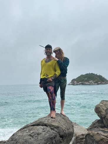 A smiling couple stands barefoot on seaside rocks. The man wears a yellow sweater and colorful leggings, while the woman embraces him. Ocean and island background.
