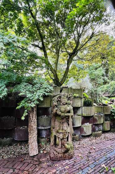 A serene garden scene features a detailed stone statue, lush green trees overhead, brick pathway, and stacked stone wall, creating a peaceful and harmonious atmosphere.