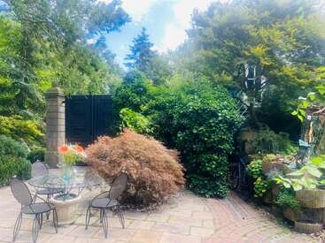 A serene garden with a glass table, four chairs, vibrant flowers, lush greenery, stone walls, a black gate, a bicycle, and sunlight filtering through trees.