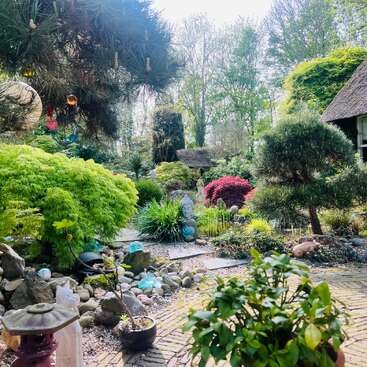 A serene garden with lush greenery, decorative rocks, a Buddha statue, vibrant plants, a thatched-roof house, and handcrafted ornaments, evoking a peaceful, Zen-like atmosphere.