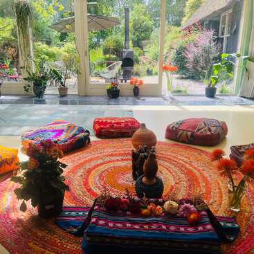 Um espaço de meditação aconchegante e colorido com almofadas estampadas, flores e um tapete redondo vibrante fica de frente para grandes janelas com vista para um jardim exuberante banhado pela luz do sol. Ambiente tranquilo.