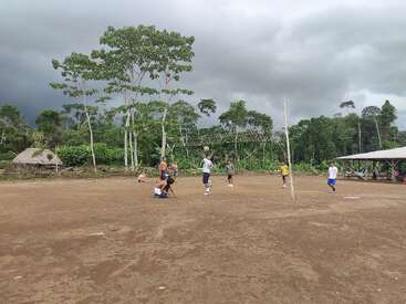 Um grupo de pessoas joga vôlei em um campo de terra batida cercado por uma vegetação exuberante, árvores altas, cabanas e um céu nublado e dramático em um ambiente rural.