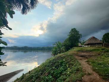 Uma cena tranquila à beira do rio ao pôr do sol, com vegetação exuberante, um caminho de terra, plantas tropicais e cabanas tradicionais com telhado de palha sob um céu dramático e cheio de nuvens. Tranquilo e remoto.