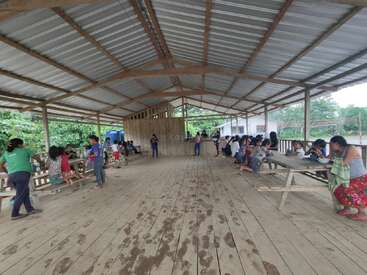 Um grande abrigo aberto de madeira com teto de metal, cheio de pessoas sentadas em bancos, comendo, conversando e socializando em um ambiente rural e verdejante.