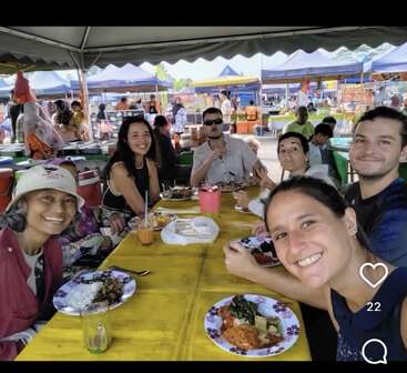 Six personnes souriantes partagent un repas sur un marché en plein air animé, assises autour d'une table jaune sous une tente, profitant de la nourriture, des boissons et de la compagnie des autres.
