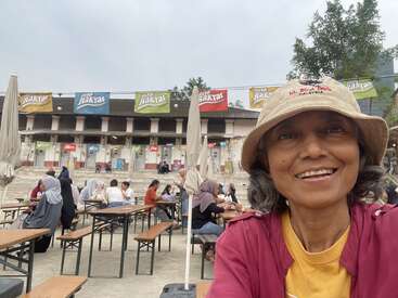 Une femme coiffée d'un chapeau sourit à l'appareil photo dans une aire de restauration en plein air. Les gens sont assis sur des bancs, avec des bannières colorées et des stands de nourriture en arrière-plan.