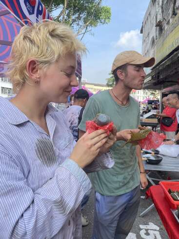 Deux personnes dégustent de la nourriture de rue sur un marché en plein air. Tous deux ont l'air heureux, savourant des en-cas traditionnels enveloppés dans du plastique et des feuilles, au milieu de l'activité grouillante du marché.