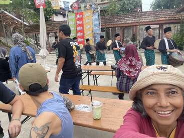 Un groupe de personnes profite d'un événement culturel en plein air. Des musiciens en tenue traditionnelle se produisent, tandis que d'autres regardent et interagissent. Une femme sourit à la caméra.