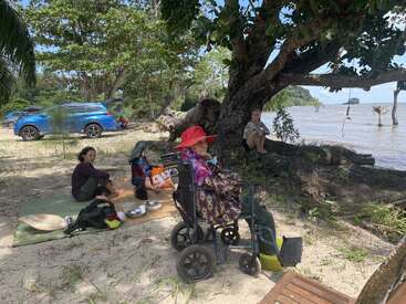 Un groupe se détend sur la plage, sous un grand arbre. Une femme âgée est assise dans un fauteuil roulant, d'autres sont assis sur des nattes, entourés par la nature et les voitures en stationnement.