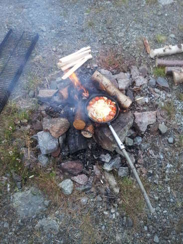 La imagen representa una hoguera con una cacerola que contiene comida, situada sobre un suelo rocoso, rodeada de troncos y una parrilla, con una rejilla metálica cerca.