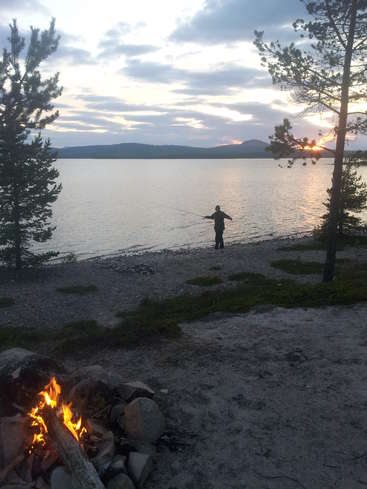 La imagen representa una serena escena junto a un lago al atardecer, con una persona pescando, una hoguera y árboles, con un ambiente tranquilo y un hermoso cielo.
