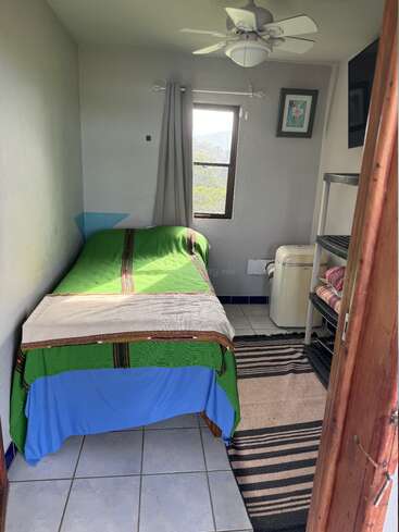 A imagem mostra um pequeno quarto com uma cama de solteiro, uma janela e um ventilador de teto, com piso de azulejos e um tapete listrado, com vista para o exterior.
