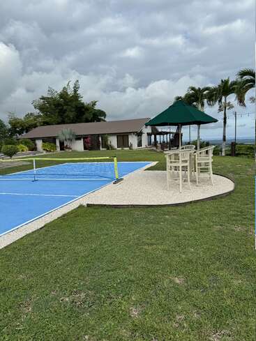A imagem mostra um ambiente externo sereno com uma quadra de tênis azul, um gazebo verde e uma casa ao fundo, em um céu nublado.